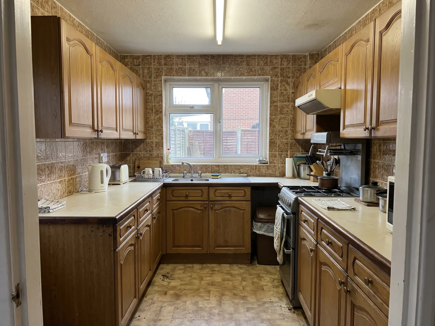 A dated kitchen with oak-effect cupboards, cream worktops, patterned brown tiles and a strip light